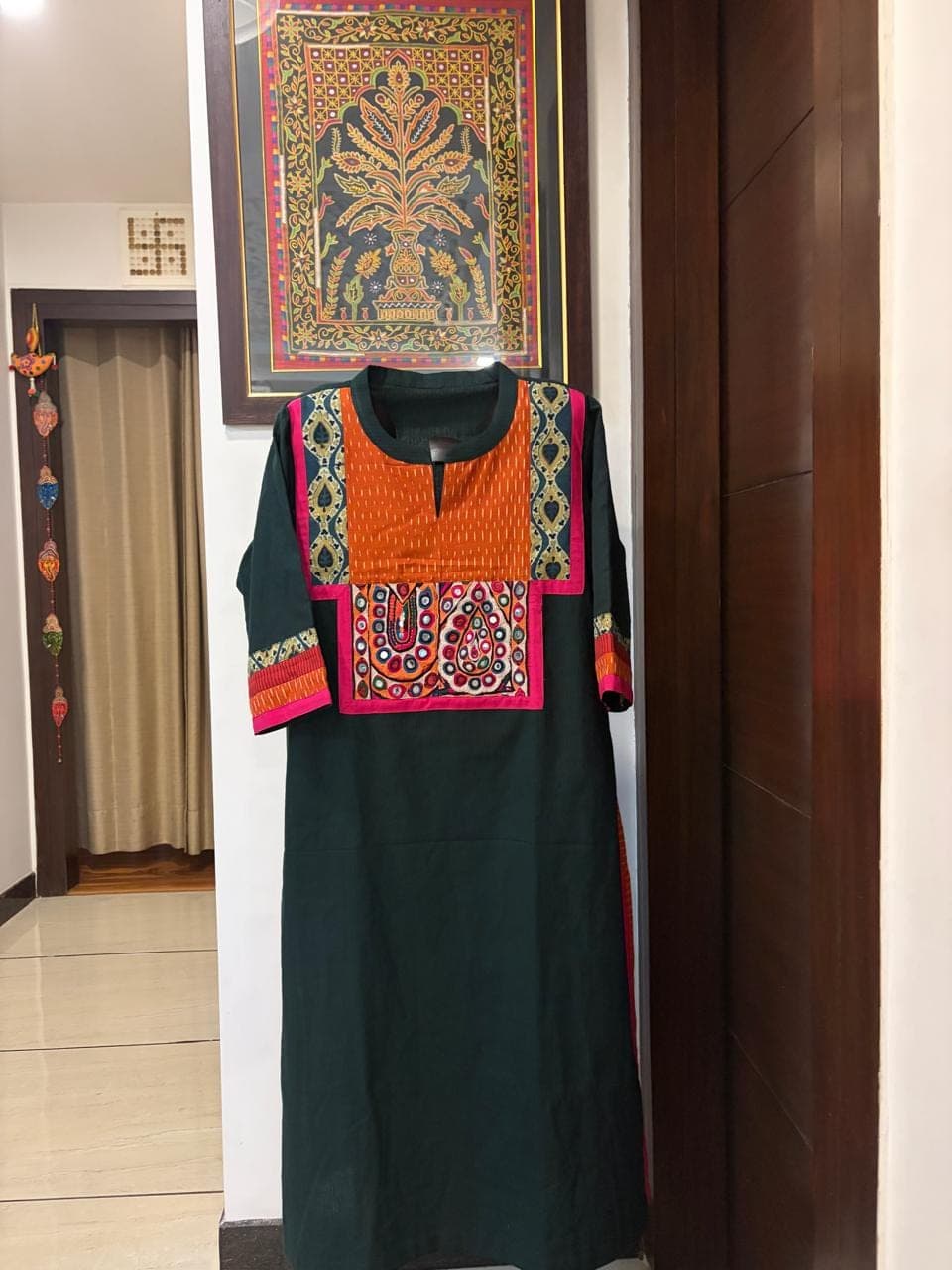 Deep green kurti with an orange mirrorwork yoke, pink piping and patterned cuffs, on a stand in a hallway