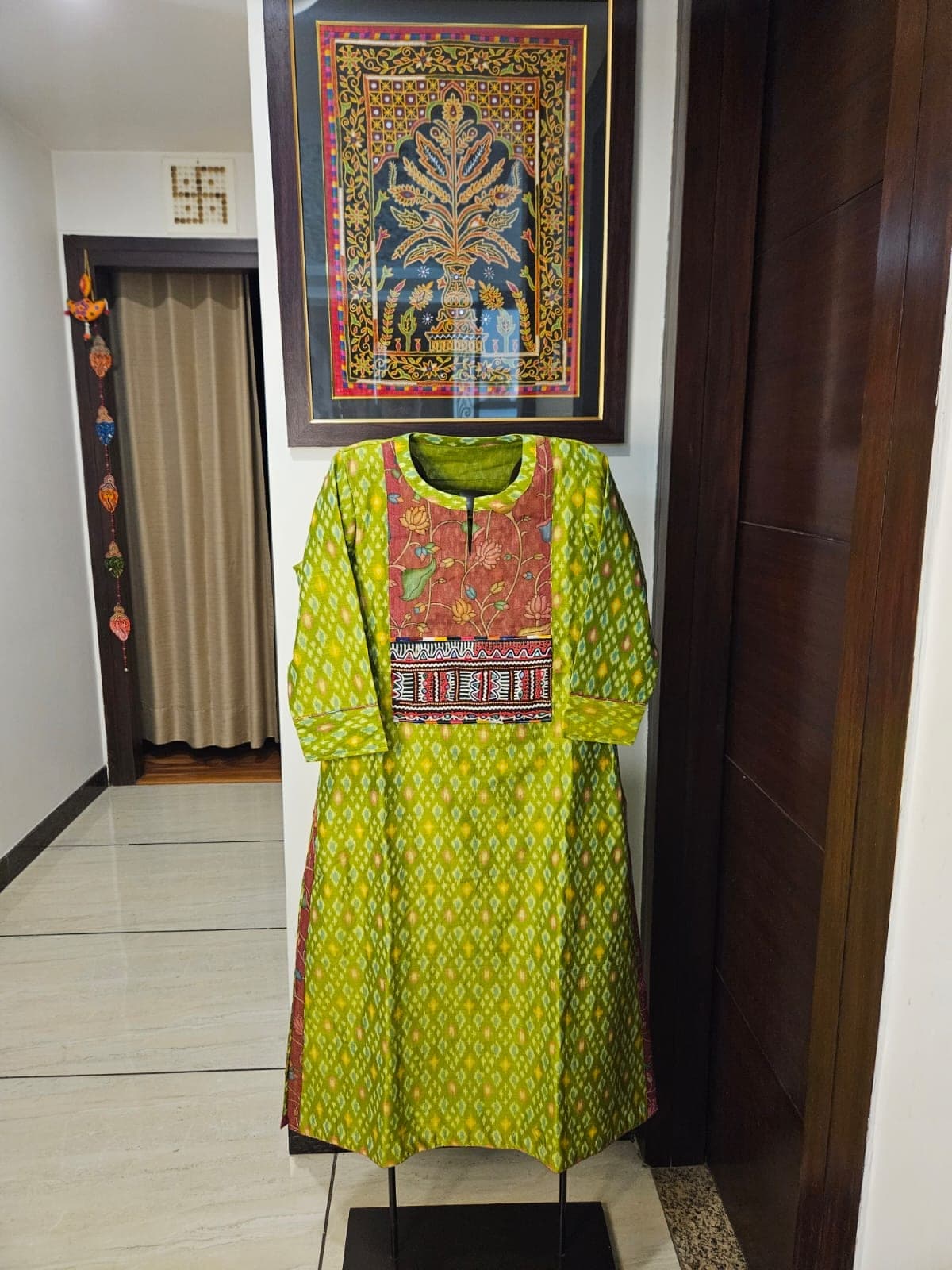 Lime green ikat kurti with a kalamkari floral yoke and an embroidered band, on a stand in a hallway