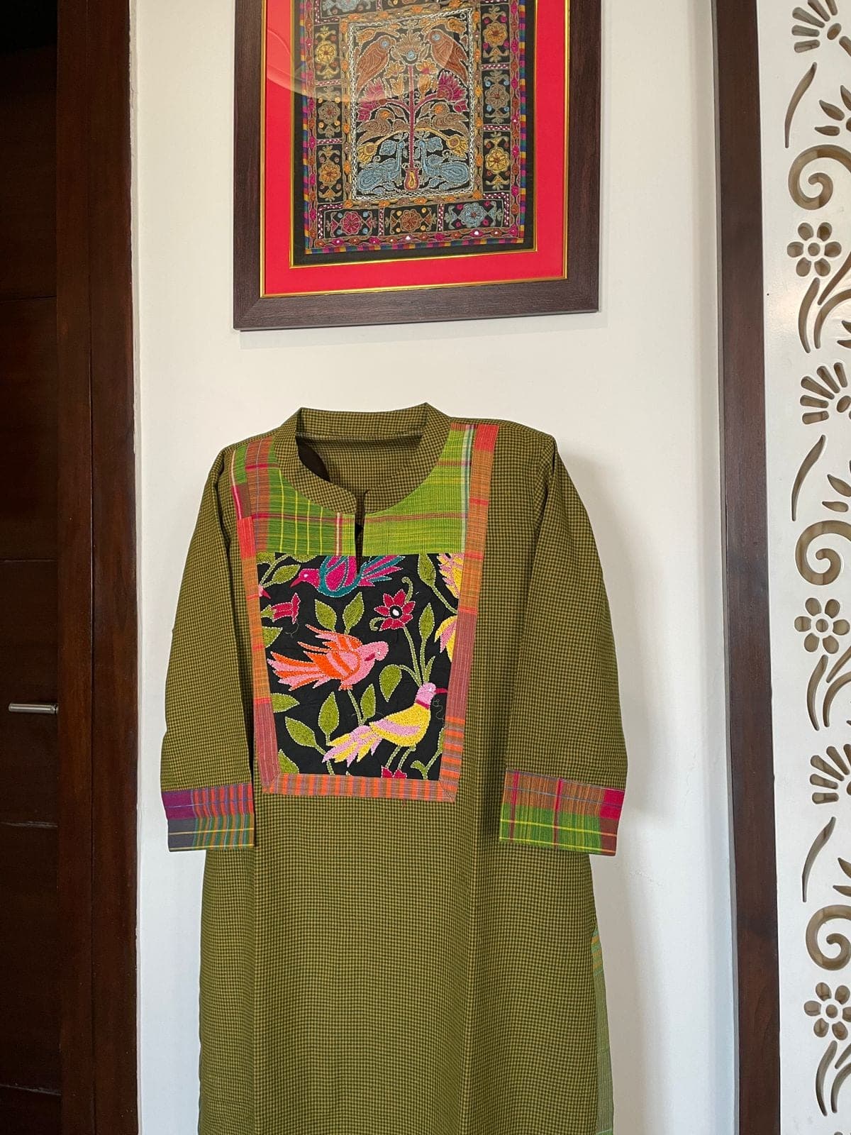 Olive checked kurti with a black panel of colourful bird and flower embroidery and checked cuffs, on a stand in a hallway