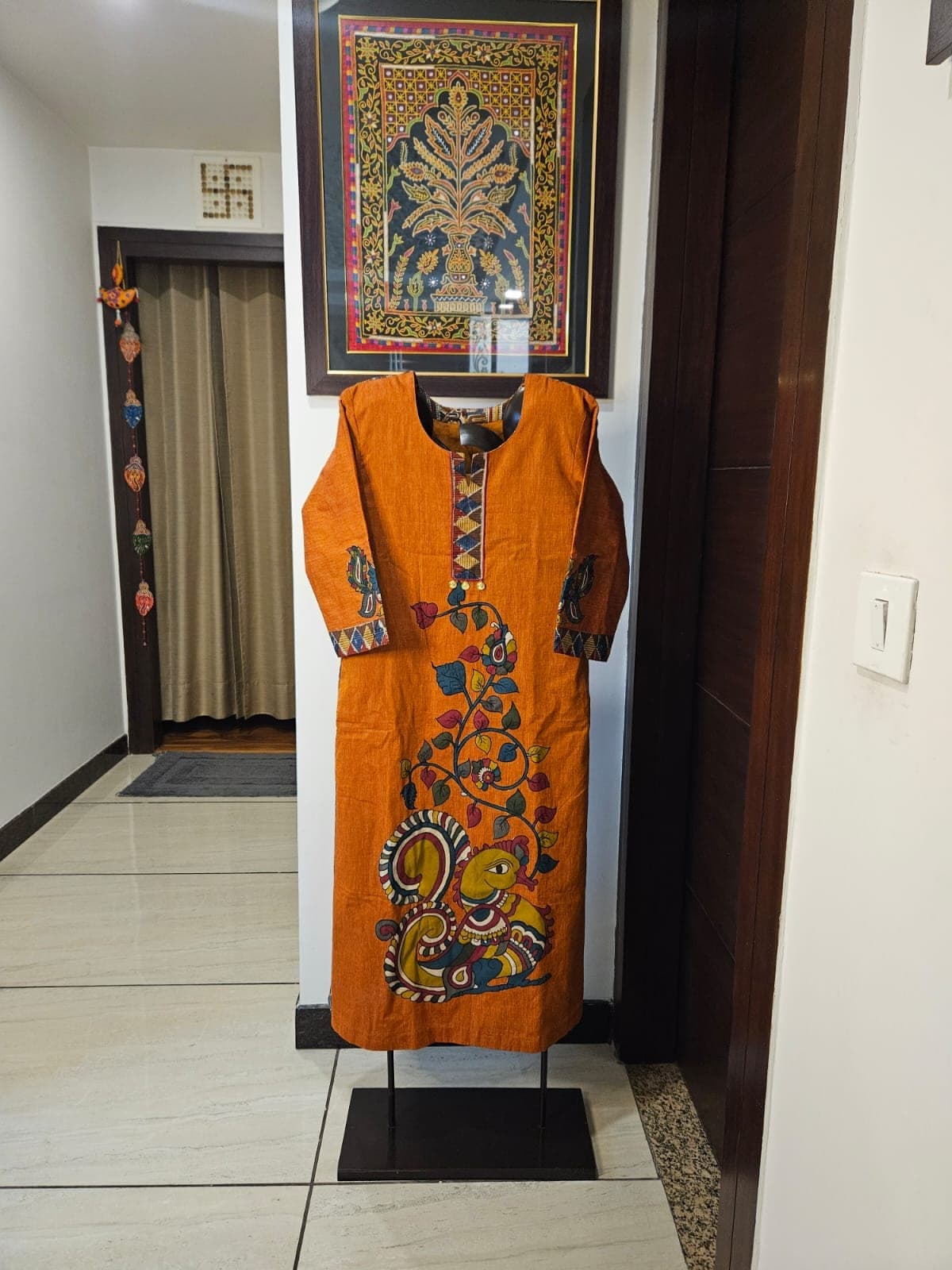 Rust kurti with a large kalamkari peacock-and-vine patchwork down the front, on a stand in a hallway
